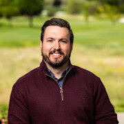 A man in a burgundy sweater smiles in front of a field.