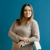 A woman with long hair, wearing a gray sweater, leans against a white chair in front of a blue wall.